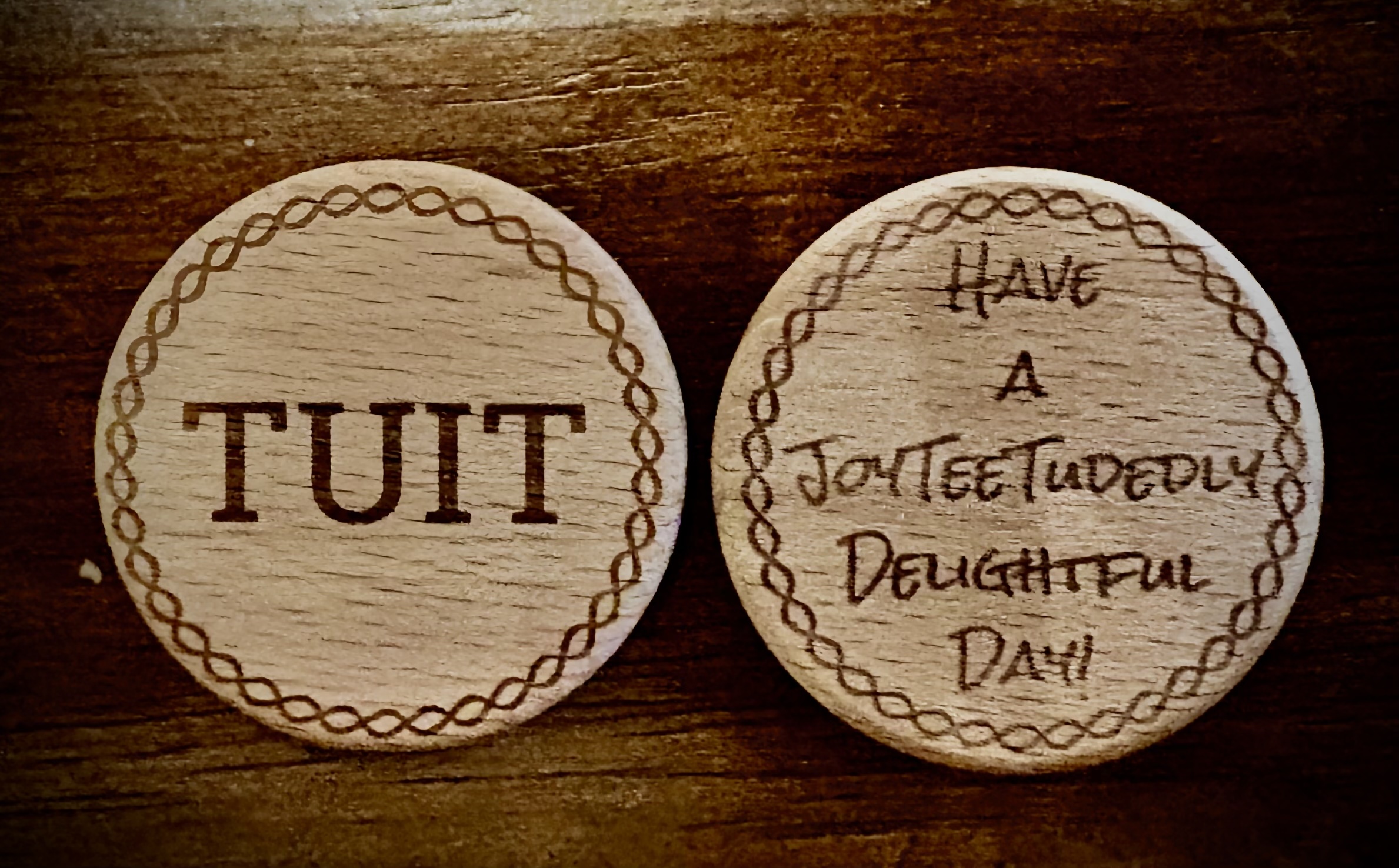 Two wooden coins on a dark table — one engraved 'TUIT', the other engraved 'Have a JoyTeeTudedly Delightful Day!'